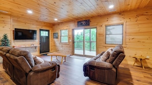 A wooden cabin interior with two reclining chairs, a flat-screen TV, a wooden bench, and a sliding glass door leading to an outdoor deck.