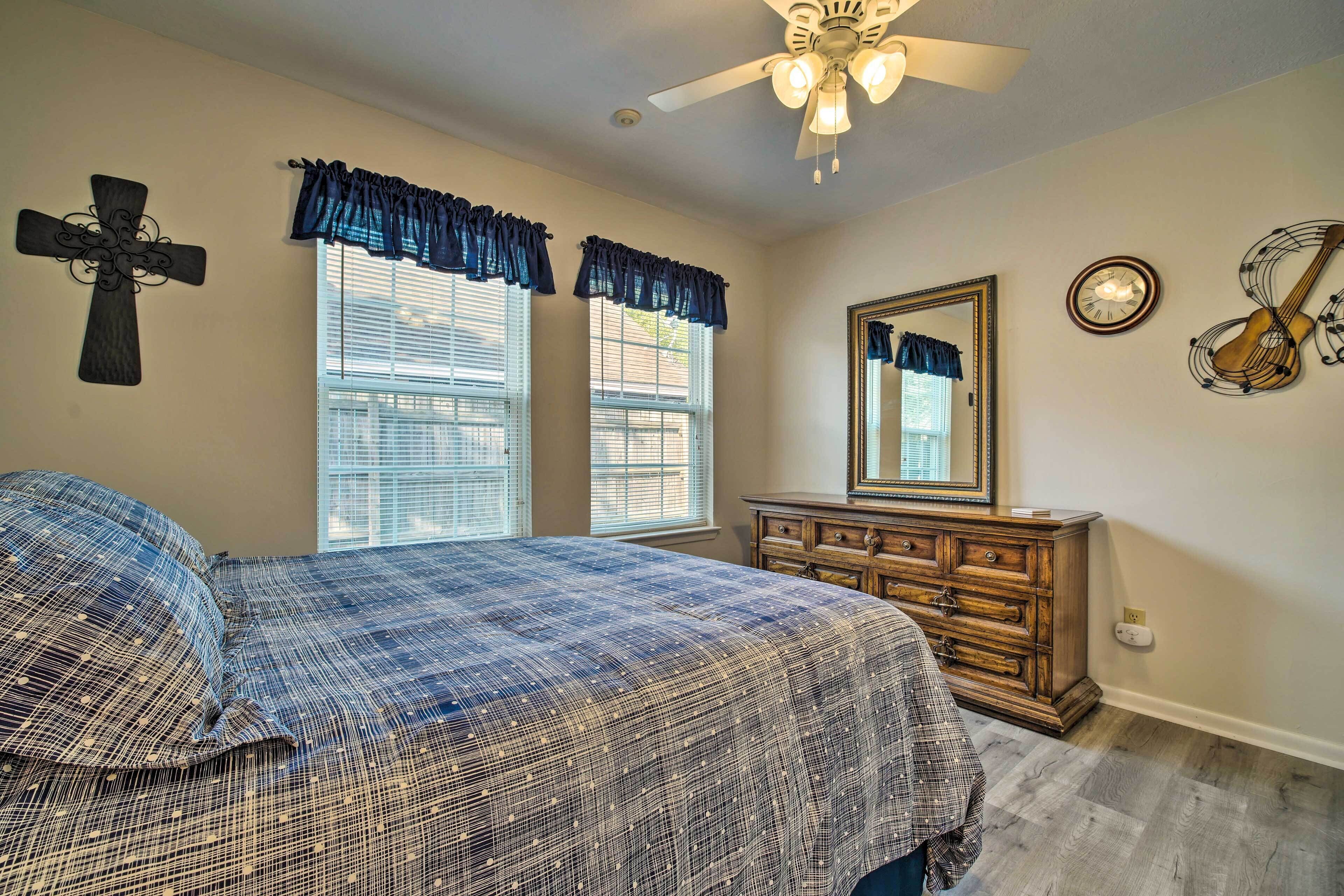A bedroom with a bed, dresser, windows with curtains, a ceiling fan, a cross on the wall, and a clock.