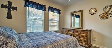 A bedroom with a bed, dresser, windows with curtains, a ceiling fan, a cross on the wall, and a clock.