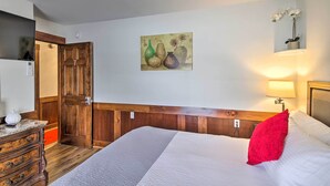 Un dormitorio con cabecera de madera, cama con ropa blanca y almohada roja, cómoda de madera y un cuadro de jarrones en la pared.