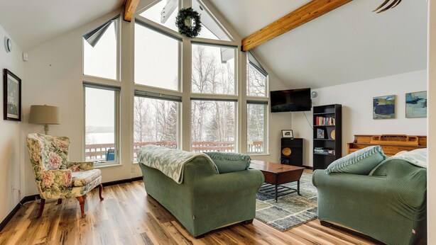 A spacious living room with a high ceiling, large windows, a wooden floor, a green sofa, a floral armchair, and a bookshelf. - Spacious and Lakefront Wasilla Home (Wasilla)