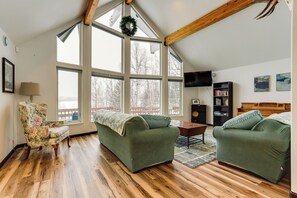 A spacious living room with a high ceiling, large windows, a wooden floor, a green sofa, a floral armchair, and a bookshelf. - Spacious and Lakefront Wasilla Home (Wasilla)
