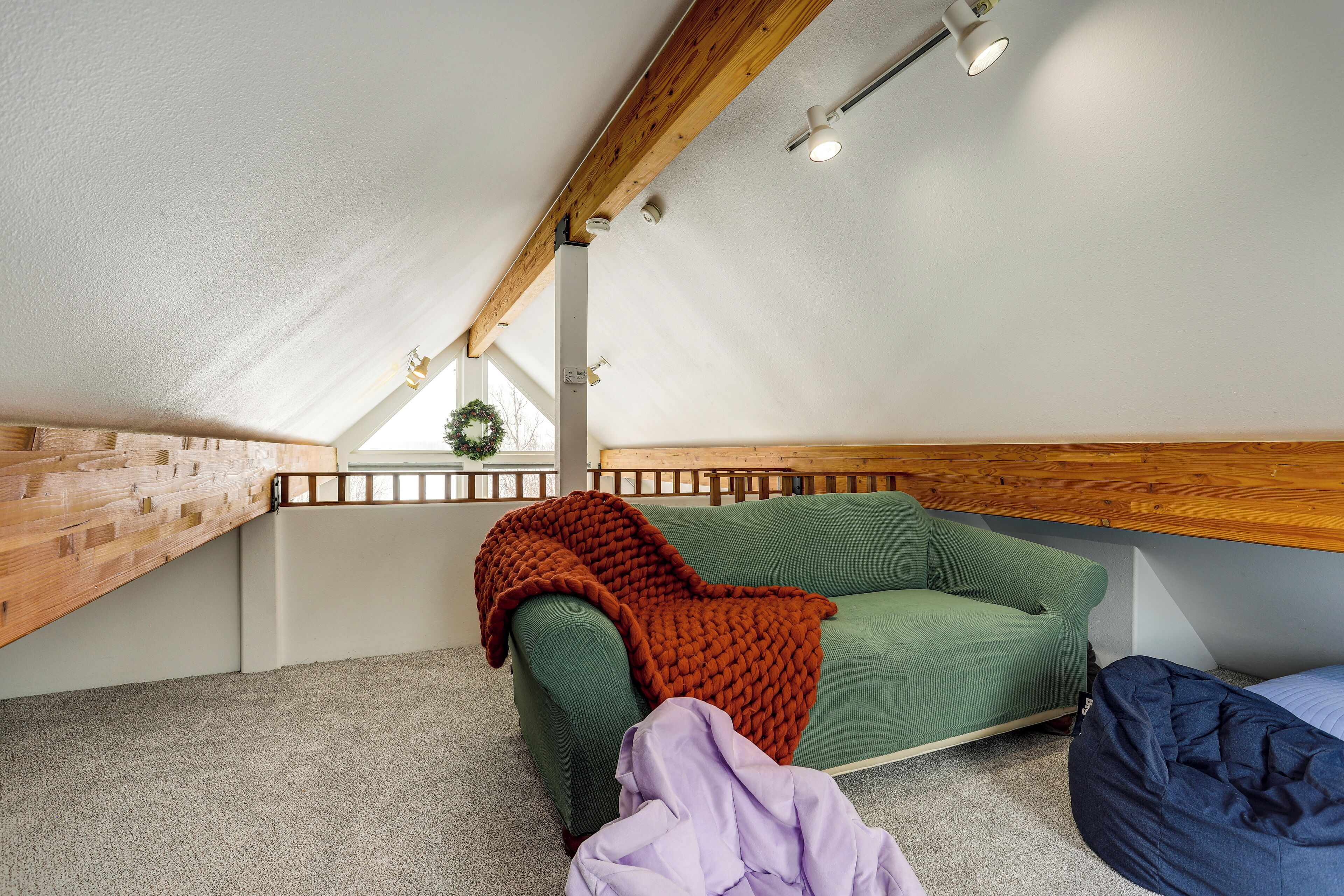 A cozy attic bedroom with a green sofa, a wooden ceiling, and a small window with a wreath.