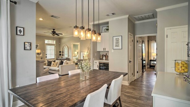 A spacious dining area with a dark wooden table, white chairs, and pendant lights. A kitchen is visible in the background with white cabinets and a window. - Baton Rouge Game Day House w/ Chic Yard Space (Baton Rouge)