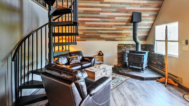 A living room with a curved staircase, a fireplace, and a stone wall. - Hillside Cripple Creek Cabin w/ Hot Tub, Mtn Views (Cripple Creek)