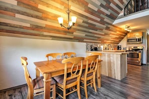 Uma sala de jantar com mesa e cadeiras de madeira, cozinha com eletrodomésticos modernos e teto inclinado com painéis de madeira.