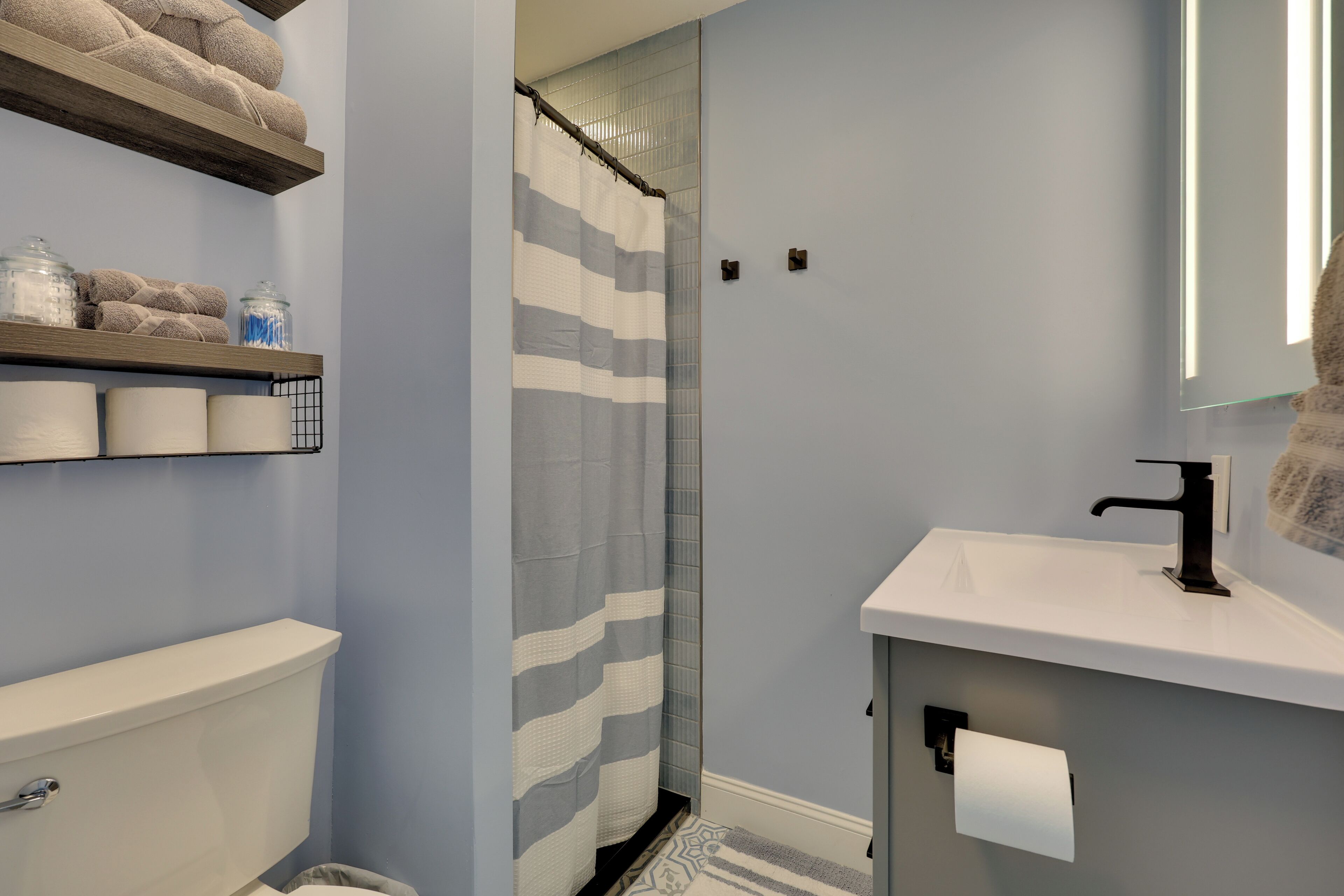 A bathroom with a toilet, a sink, a shower with a striped curtain, and shelves with towels and toiletries.