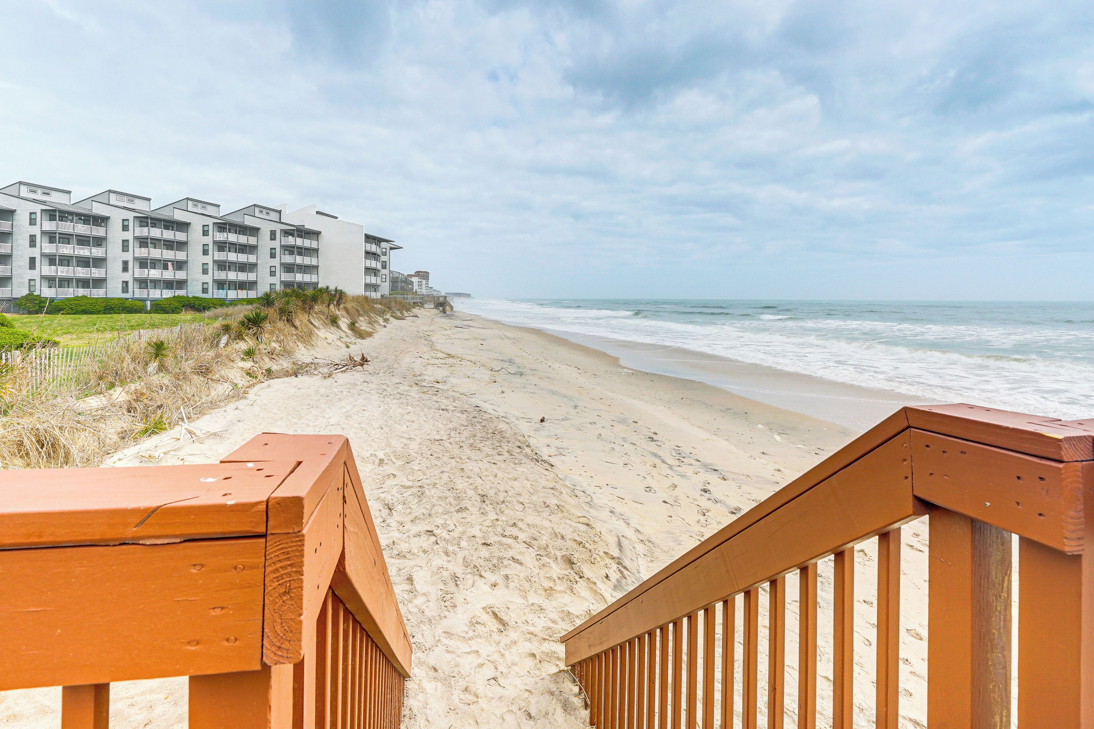 A beachside view with a wooden walkway leading to a building with multiple balconies.