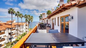 A balcony with a table and chairs overlooking a coastal area with palm trees and a view of the sea.