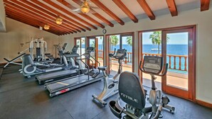 A gym with various exercise equipment, large windows with a view of the ocean, and wooden ceiling beams.