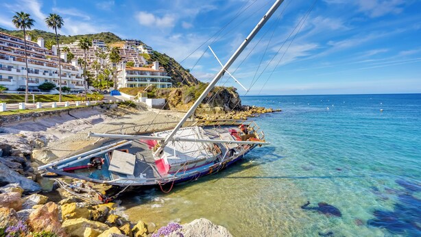 A coastal scene with a boat stranded on rocks, clear blue water, and a hillside with buildings.