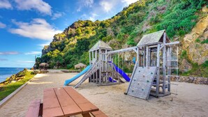 A beach playground with slides and climbing structures, a picnic table, and a view of the ocean.
