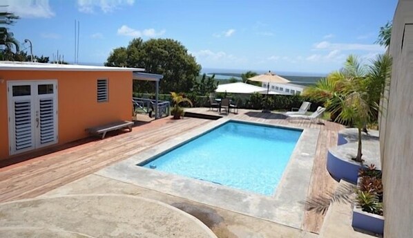 A rectangular swimming pool with a wooden deck, a small orange building with shutters, and a view of the ocean.