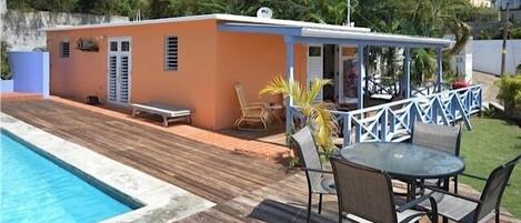 A poolside area with a wooden deck, a small building with a white roof, and outdoor furniture.