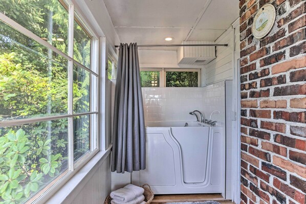 A bathroom with a bathtub, a shower, and a window with curtains. - Charming Historic Retreat Near Poverty Bay! (Federal Way)