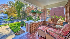 A covered porch with a brick wall, a brick fireplace, a sofa, and a dining table. - Vintage Chic Cottage < 1 Mile to UNC Greensboro! (Greensboro)
