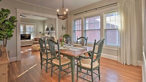 A dining area with a wooden table, green chairs, and a chandelier. There are windows with curtains and a brick wall visible in the background. - Vintage Chic Cottage < 1 Mile to UNC Greensboro! (Greensboro)