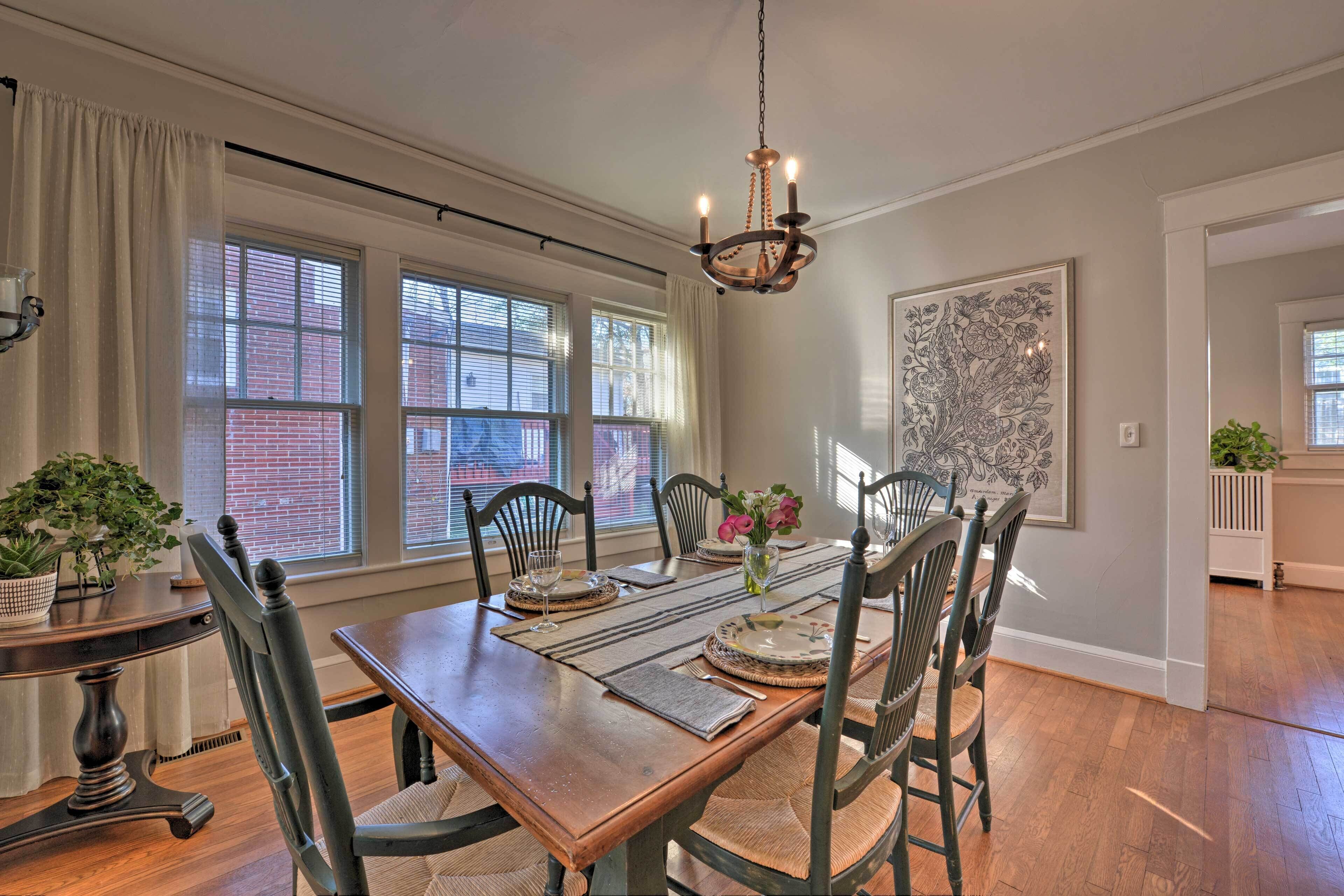 A dining room with a wooden table set for four, surrounded by chairs. There are large windows with blinds, a chandelier, and a framed artwork on the wall.
