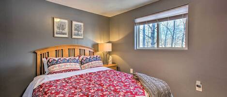 Ein Schlafzimmer mit einem Holzkopfende, einer rot-blumigen Tagesdecke, einem Fenster mit Jalousien und zwei gerahmten Bildern an der Wand.