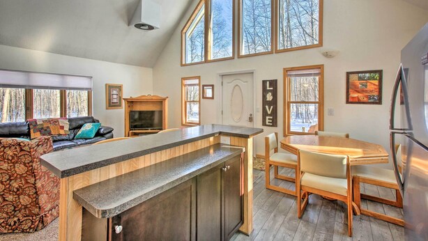 Una cocina moderna con una isla central, una mesa de comedor de madera y sillas. Se observa un horno empotrado y un refrigerador a la derecha. La cocina tiene ventanas amplias y vistas a árboles.