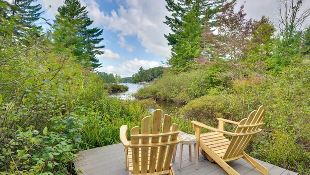 Eine Holzterrasse mit zwei Adirondack-Stühlen und einem kleinen Tisch, mit Blick auf einen Fluss und dichtes Grün.