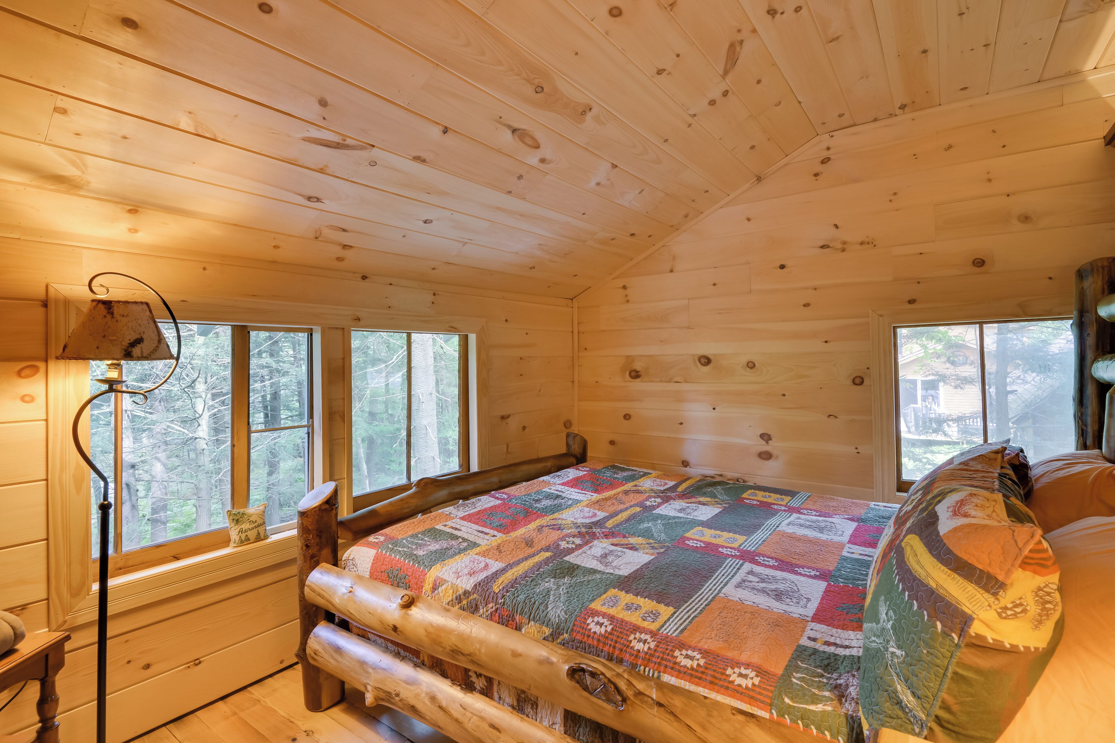 A cozy cabin room with a wooden bed, a quilt, a lamp, and large windows offering a view of the forest.