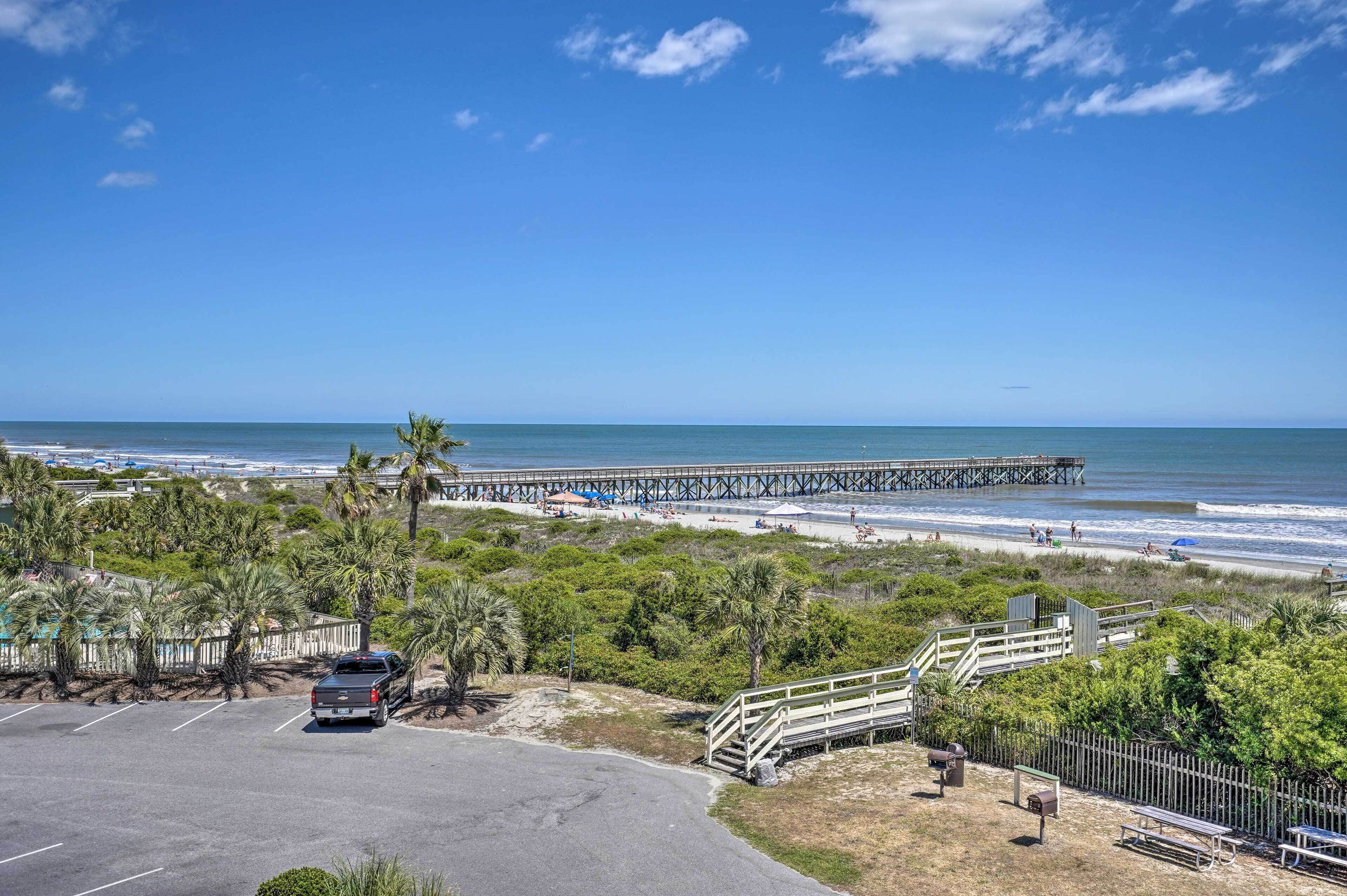 A coastal view with a pier extending into the ocean, a parking area, and a beach with people and umbrellas.