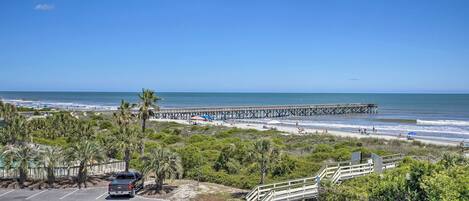 A coastal view with a pier extending into the ocean, a parking area, and a beach with people and umbrellas.