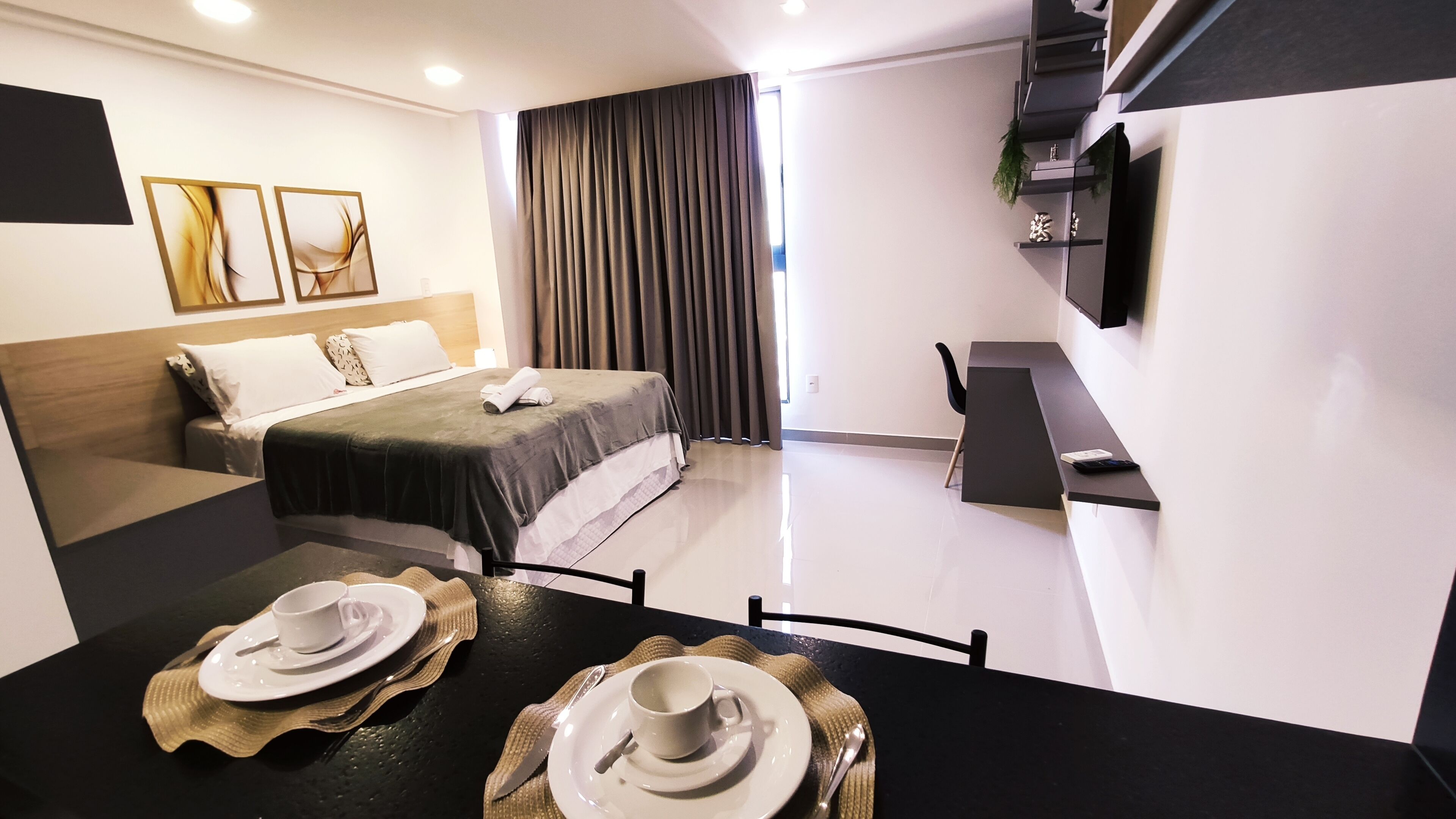 Ein modernes Hotelzimmer mit einem ordentlich bezogenen Bett, einem Esstisch gedeckt mit Tellern und Tassen, einem an der Wand montierten Fernseher und einem großen Fenster mit Vorhängen.