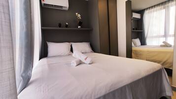 A hotel room with a bed, a bedside table, a small shelf with decorative items, and an air conditioning unit.