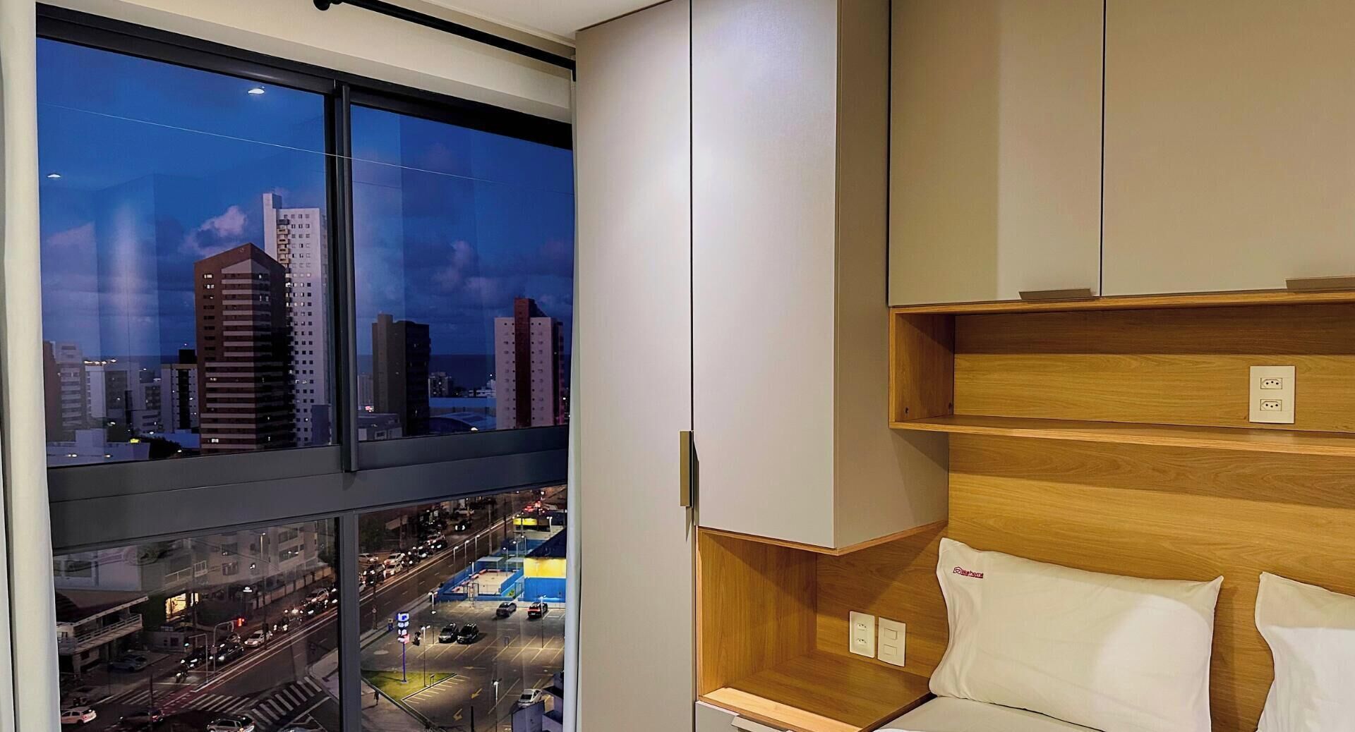 A modern hotel room with a city view, built-in shelving, and a bed with a pillow.