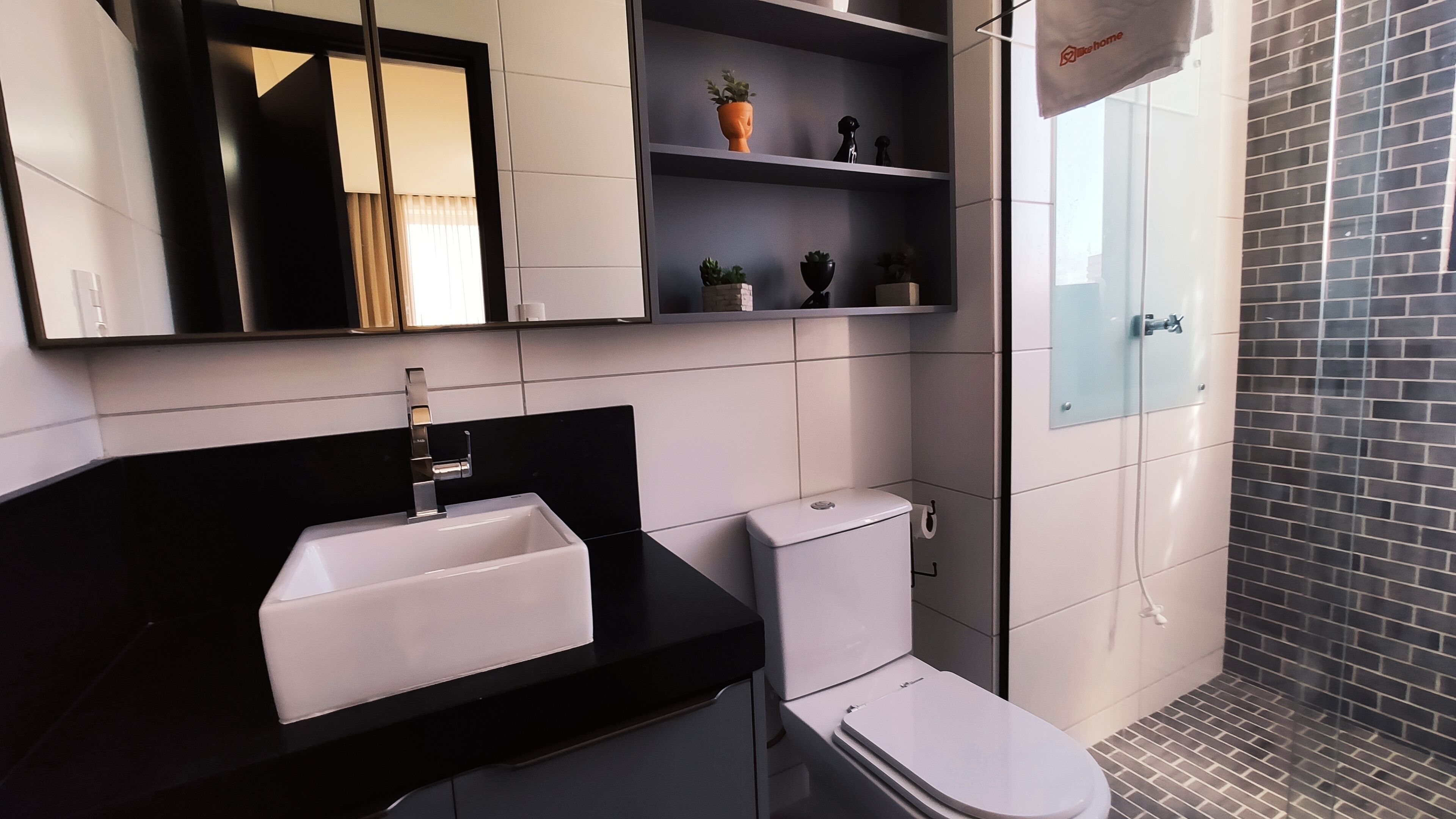 Un baño moderno con encimera negra, lavamanos blanco y retrete. Hay una repisa con objetos decorativos y una toalla colgada en la pared.