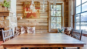A rustic dining area with a wooden table and benches, a stone wall, and a large window with a view of trees. - 2 Blocks To Grand Lake: Downtown Western Cabin! (Grand Lake)