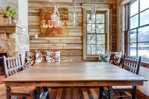A rustic dining area with a wooden table and benches, a stone wall, and a large window with a view of trees.
