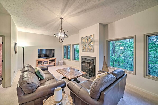 A living room with a fireplace, leather sofas, a coffee table, and large windows offering a view of trees. - Wintergreen Resort Home w/ Deck! (Roseland)