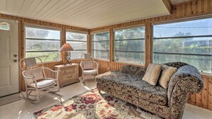 A room with a wooden ceiling, large windows, a patterned sofa, a floral rug, and a wicker chair. - Charming Port Charlotte Home w/ Lanai & Pool! (Port Charlotte)