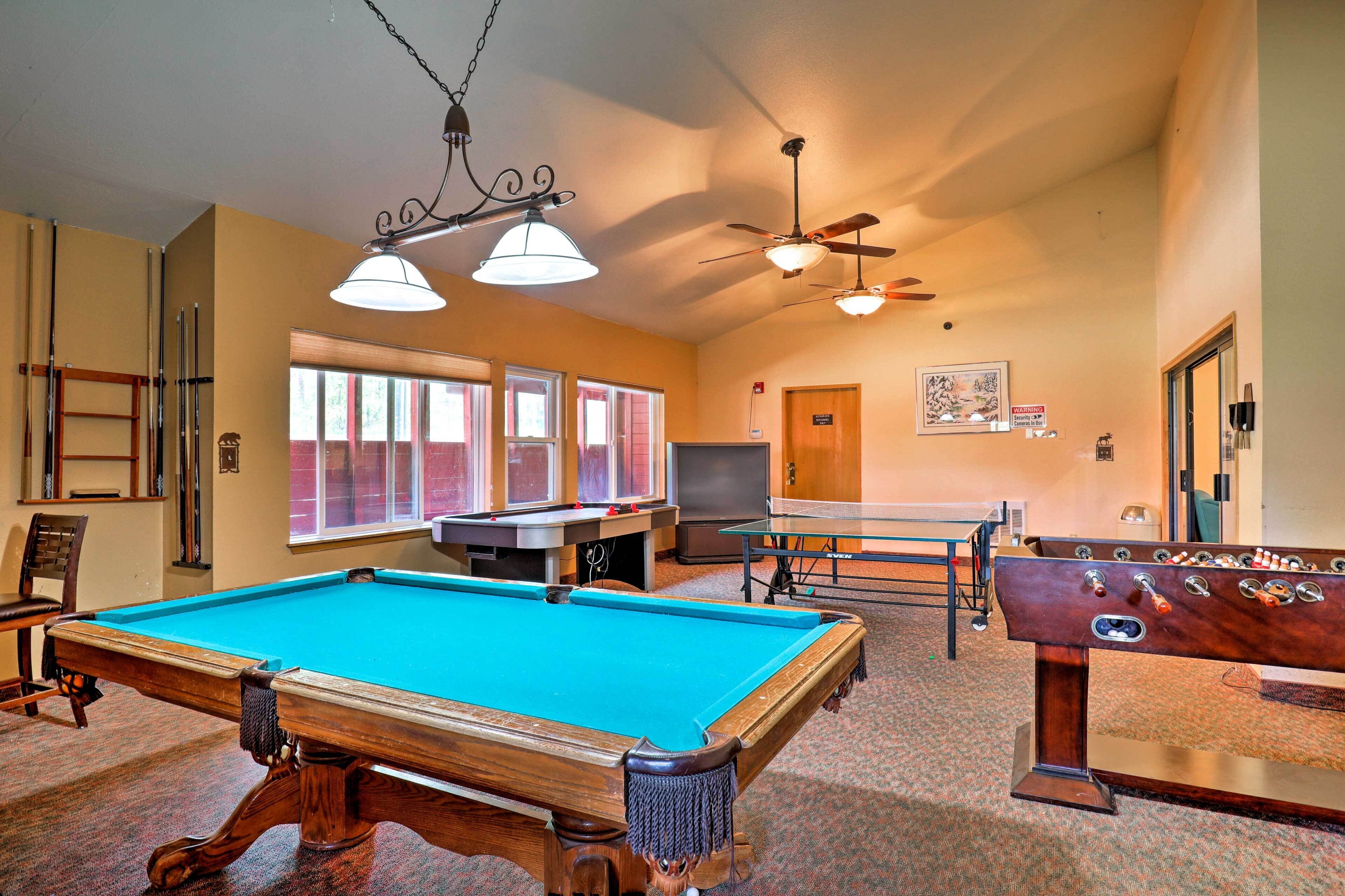 A pool table, foosball table, ping pong table, and a billiards table in a room with windows and ceiling fans.