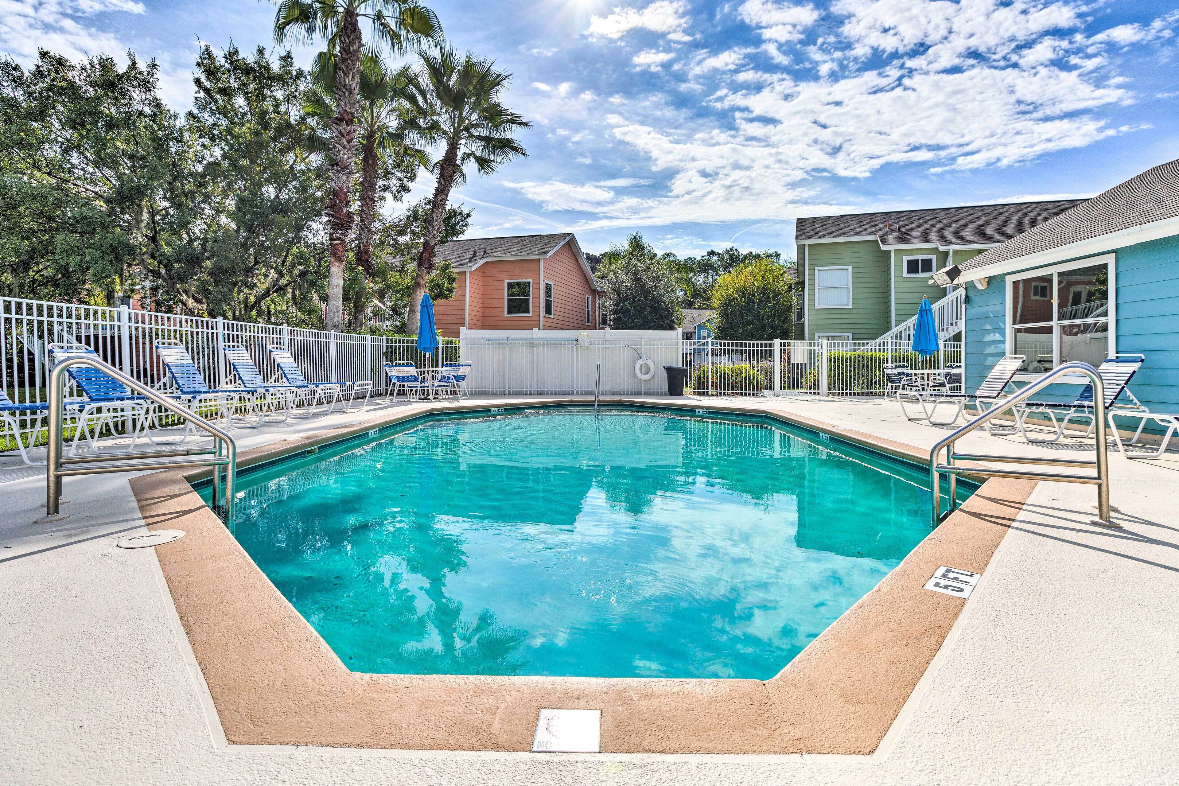 Une piscine avec des chaises longues, un espace délimité par une clôture et des maisons en arrière-plan.