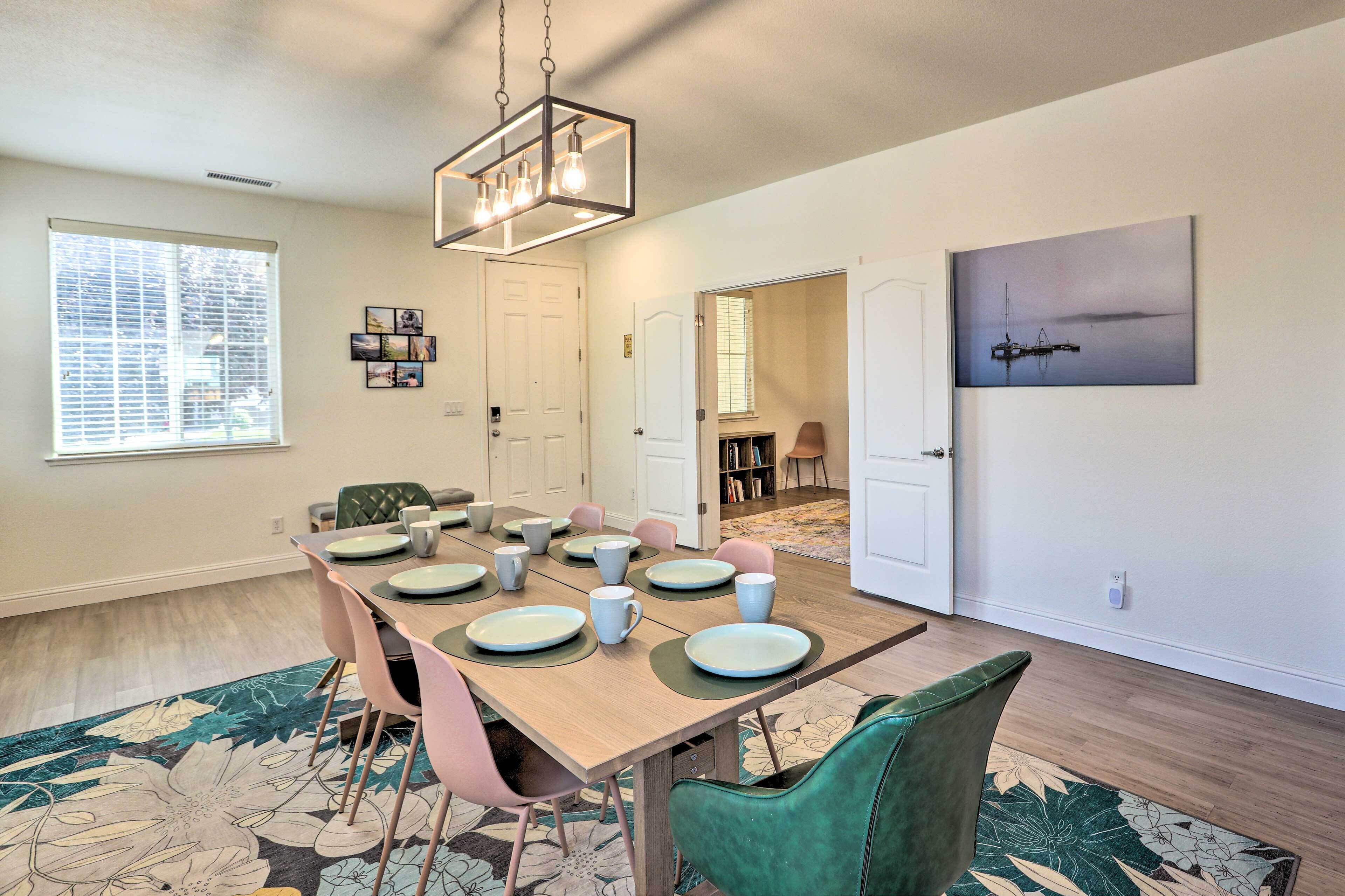 Un coin repas avec une table en bois dressée pour six personnes, entourée de chaises aux couleurs variées. On y trouve une grande fenêtre avec des stores, un luminaire au plafond et une œuvre d’art encadrée accrochée au mur.
