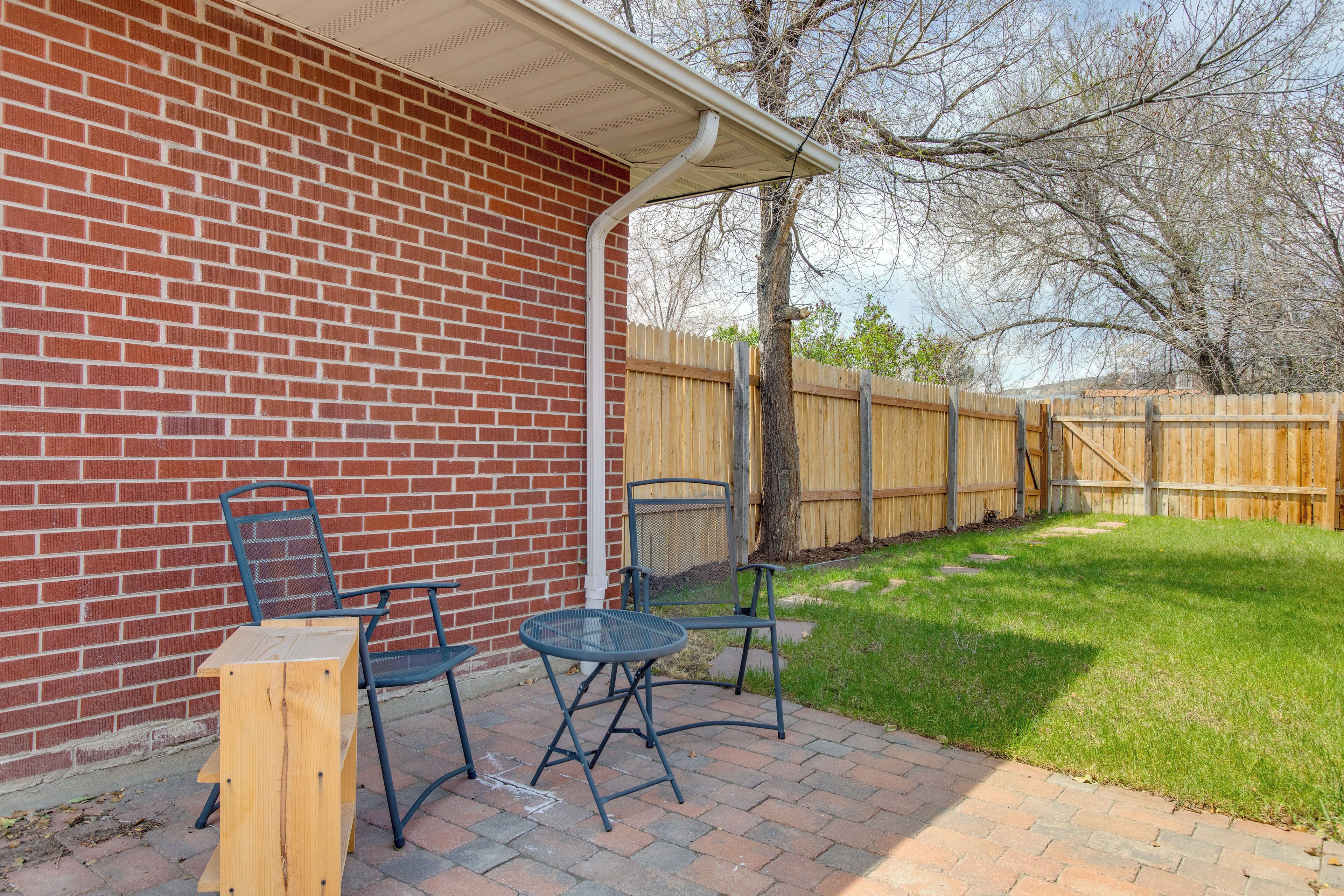 A patio with a brick wall, two metal chairs, a small table, and a wooden stool.