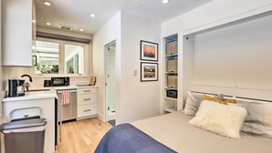 Un dormitorio moderno con una cama, un pequeño rincón americano y un baño visible a través de una puerta abierta.