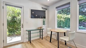 Una habitación moderna con una mesa de vidrio, sillas blancas, un televisor de pantalla plana y ventanas amplias que ofrecen vistas a zonas verdes.