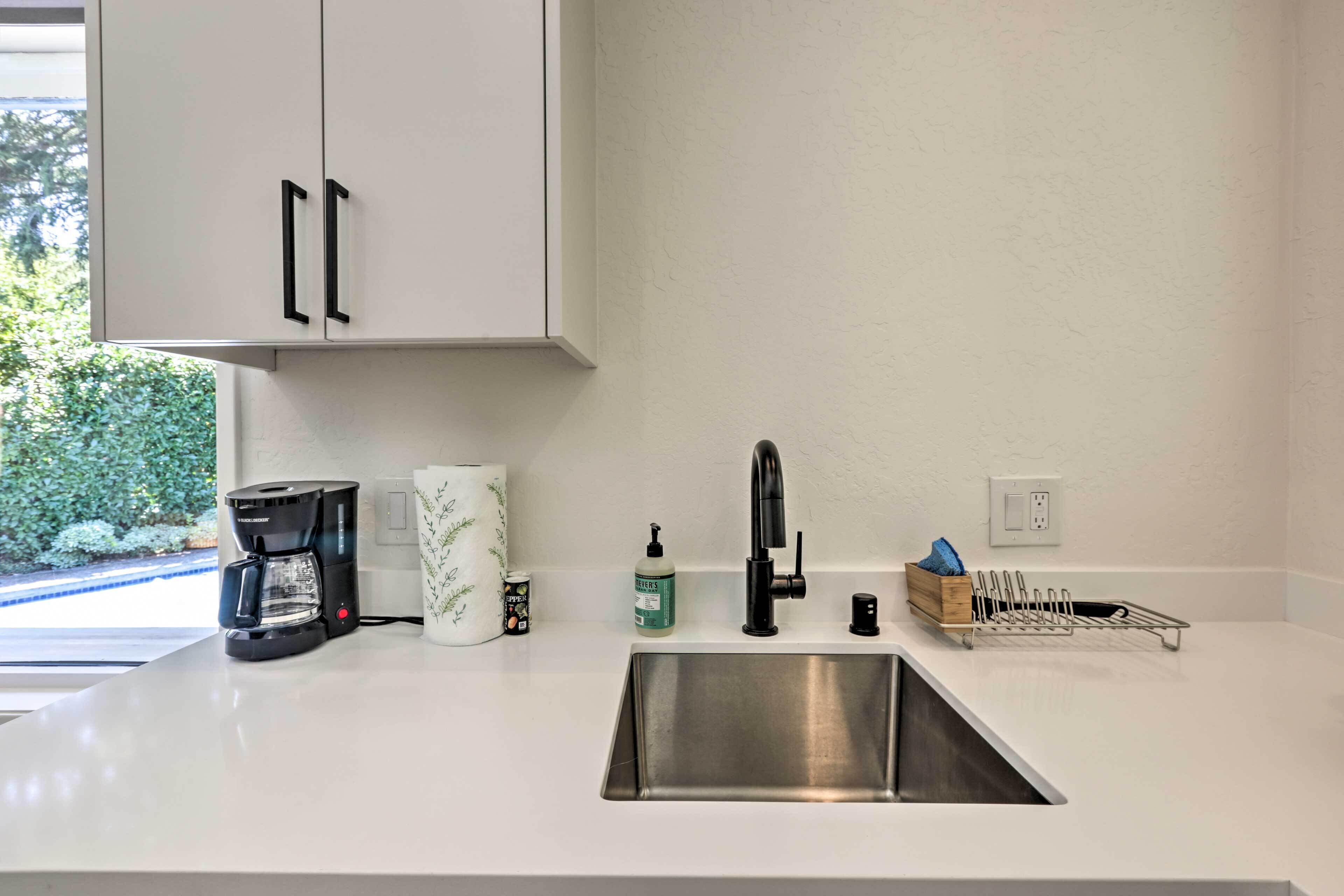 A kitchen with a stainless steel sink, a coffee maker, a soap dispenser, and a dish rack.