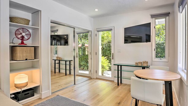 Un interior moderno con suelo de madera, un comedor con mesa y sillas redondas, un televisor montado en la pared y un amplio ventanal con vistas a la vegetación.