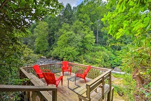 A wooden deck with red chairs overlooking a forested area and a small waterfall. - Quiet Riverfront Escape With Deck & Fly Fishing! (Sapphire)