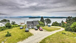 A residential area with a gravel driveway, parked cars, and houses near a lake with mountains in the background. - Acadia Home With Incredible Frenchman Bay View! (Ellsworth)