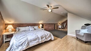 A bedroom with a sloped ceiling, a bed, a nightstand with a lamp, a grey armchair, and a brick wall. - Acadia Home With Incredible Frenchman Bay View! (Ellsworth)