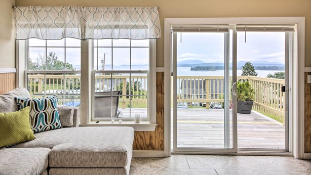 A room with a large sliding glass door leading to a deck, a cushioned bench, and a view of a deck with a railing and greenery. - Acadia Home With Incredible Frenchman Bay View! (Ellsworth)