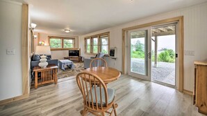 A spacious living area with a wooden dining table, chairs, and a sofa. There is a television and a small table with decorative items. Large windows allow natural light and provide views of the outdoors. - Acadia Home With Incredible Frenchman Bay View! (Ellsworth)