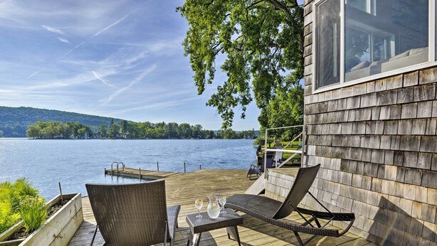Eine Holzterrasse mit Korbstühlen und einem Tisch, mit Blick auf einen See, eine Anlegestelle und sanfte Hügel in der Ferne.